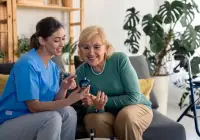 A nurse is cheerfully assisting an elderly woman in learning how to use a medical device, likely a blood glucose monitor, in a comfortable home setting.&nbsp;