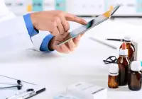 Doctor using a digital tablet in a clinical setting, surrounded by medication bottles and a stethoscope.&nbsp;