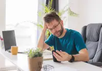 Unwell man using smartphone at home for telehealth consultation while sitting at desk.