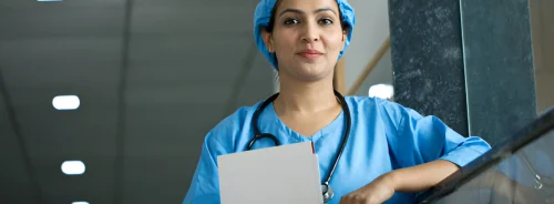 A healthcare professional in surgical scrubs and cap stands in a hospital corridor holding a clipboard, appearing ready for clinical duties.