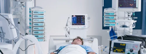 Intensive care unit scene showing a patient in bed connected to monitoring and life-support equipment.