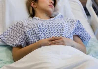 Young female patient resting in hospital bed wearing a medical gown after surgery or treatment. 