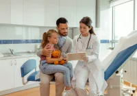 Paediatric consult: doctor reviews information on a tablet with a father and child holding a toy in a clinic. 