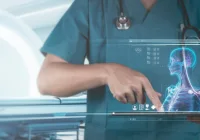 Clinician holding a tablet displaying a digital anatomical model beside an MRI scanner, illustrating integration of imaging systems with real-time data visualisation and AI-supported diagnostic workflows.