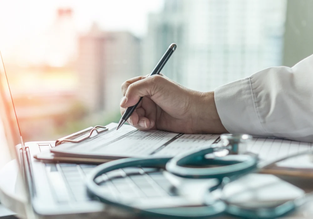 Close-up of a doctor filling out a medical form on a clipboard beside a stethoscope and laptop, representing patient documentation, healthcare compliance and clinical workflow. 