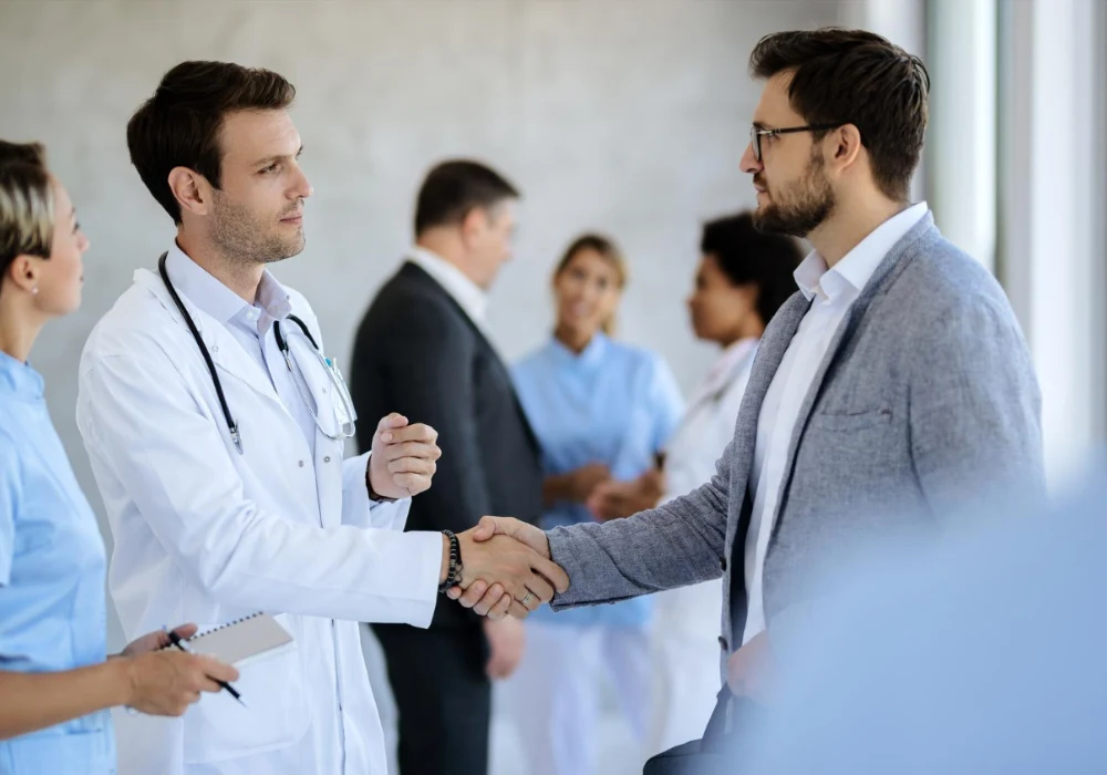 Doctor and hospital executive shake hands during merger planning meeting with clinical team. 