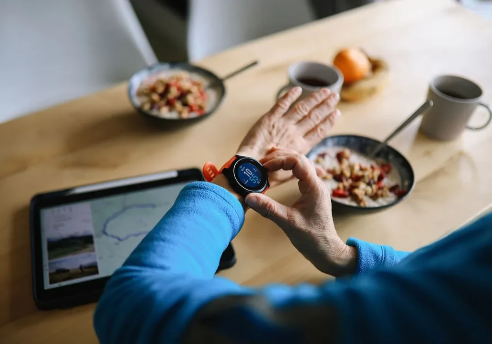 Person checking smartwatch fitness data during breakfast, next to tablet showing route map. 