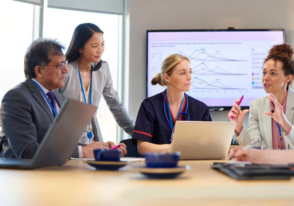 Multidisciplinary healthcare team meeting, reviewing performance data on a screen. 