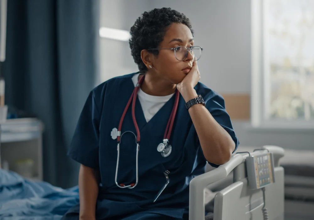 Tired healthcare worker sitting beside hospital bed, reflecting stress and frontline fatigue.