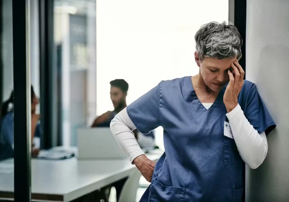 A healthcare worker in scrubs leans against a wall, holding their head, suggesting fatigue or stress in a clinical workplace.