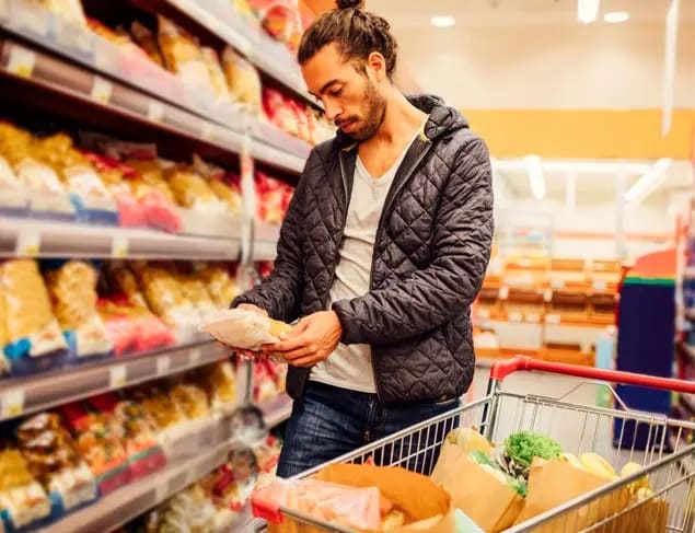 Young bearded man doing grocery shopping. He is choosing pasta and reading the nutrition label on the product