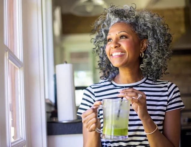 Woman with curly grey hair in a black and white striped tshirt sipping a green smoothie and looking out the window