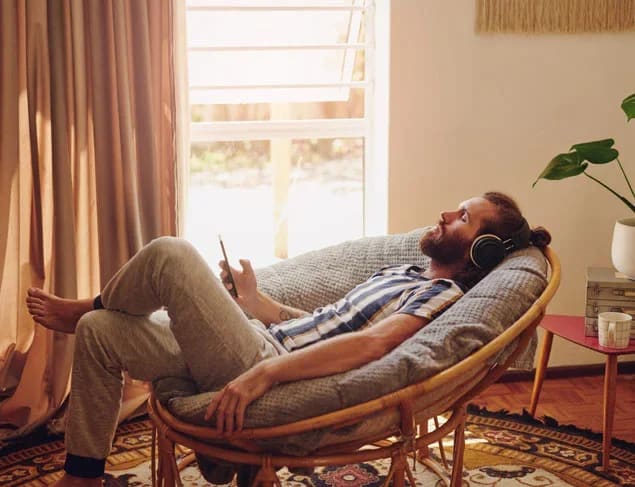 Man relaxing in a cane armchair eyes closed, wearing headphones and listening to music