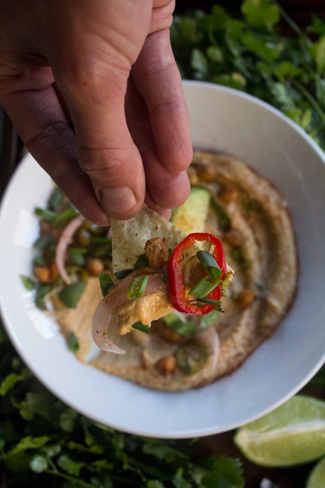 2022_TacoHummus_21web Close-up of hand holding tortilla chip and Taco Hummus with pickled salsa, crispy chickpeas, cilantro, and avocado