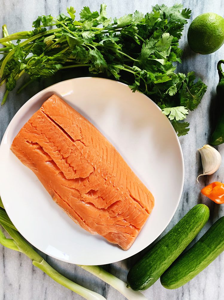 SalmonBurgers_Ingredients_web Fresh salmon on a plate with cilantro, lime, jalapeno, cucumbers, garlic, and green onions