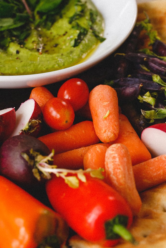 Green Goddess Hummus in shallow bowl with radishes, cherry tomatoes, flatbreads, and spring peas, sweet peppers