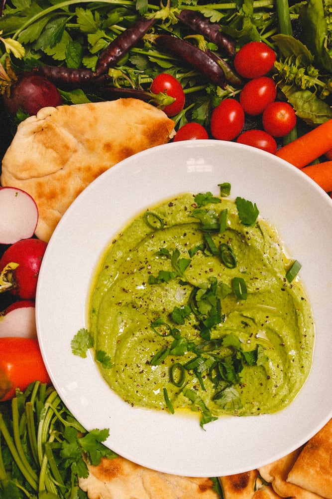 Green Goddess Hummus in shallow bowl with radishes, cherry tomatoes, flatbreads, and spring peas