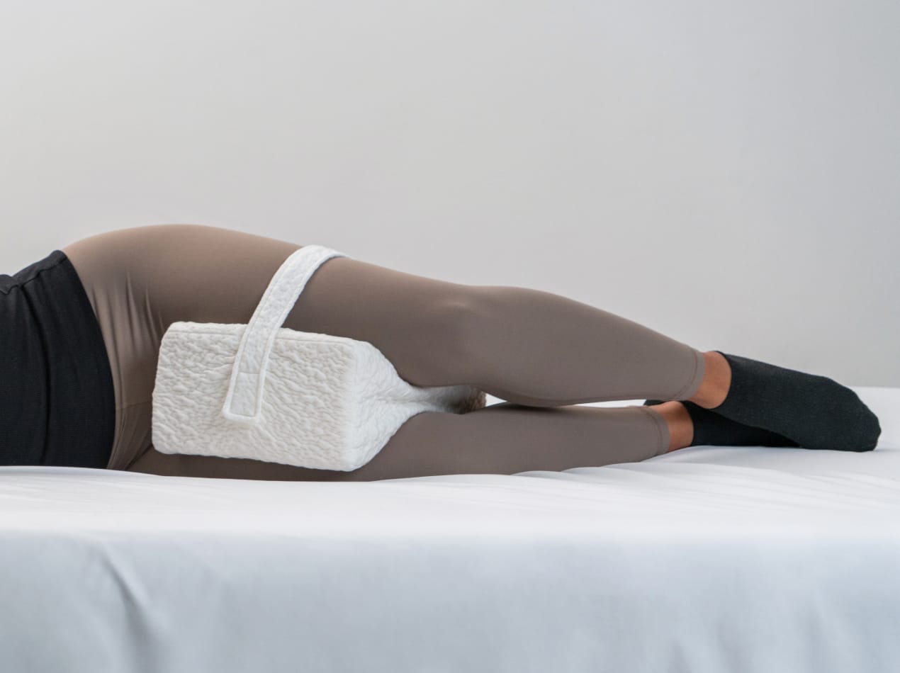 A side view of a person lying in bed, using a white contoured knee pillow with a strap to keep their legs aligned and their hips properly supported.