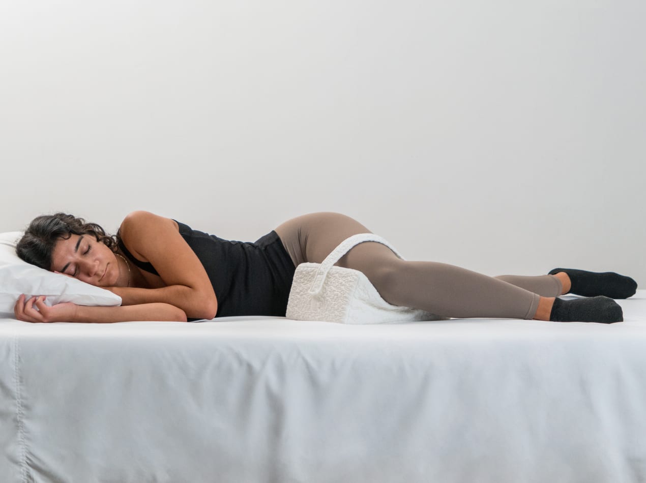 A side view of a person lying in bed, using a white contoured knee pillow with a strap to keep their legs aligned and their hips properly supported.
