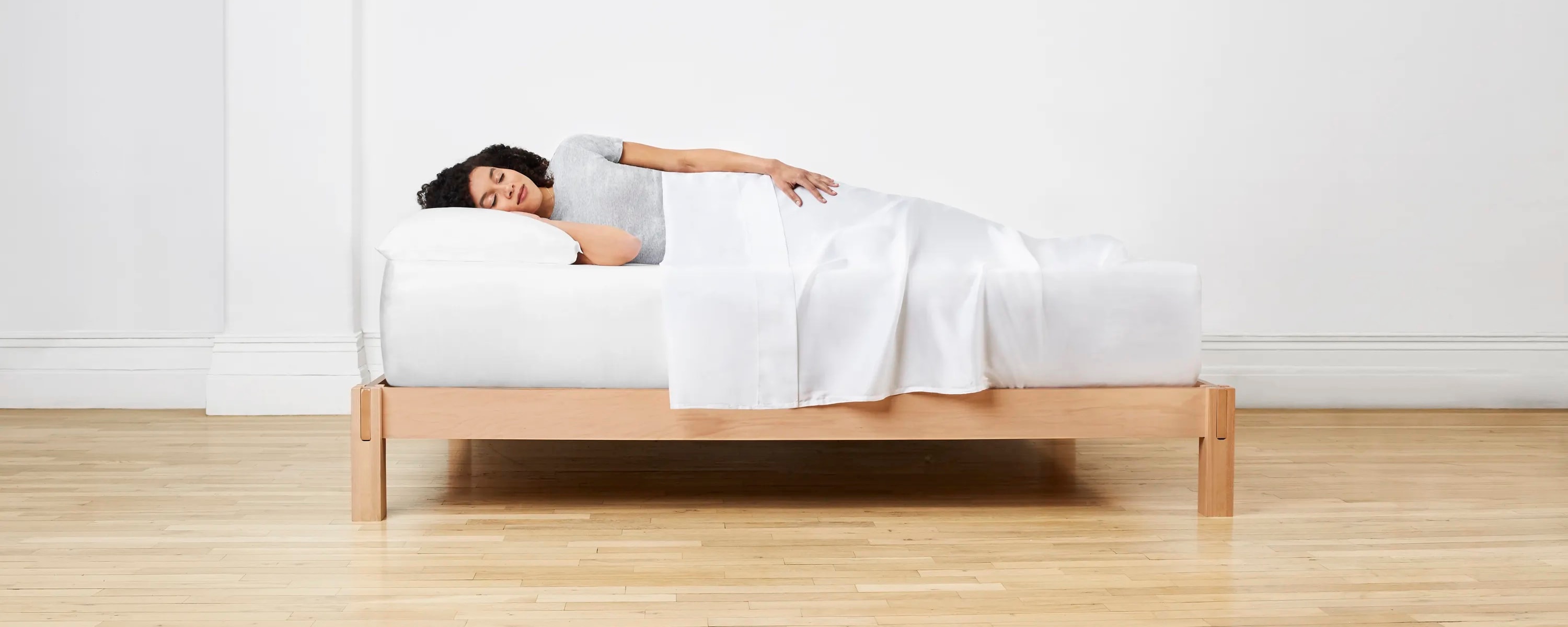 Woman laying on her side on Standard Mattress, with a bedsheet over her.  Mattress is set up on natural wood frame.