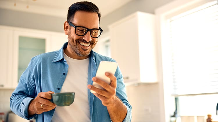 Hombre con una camisa azul, sonriendo mientras mira su teléfono, sosteniendo una taza en una cocina luminosa.