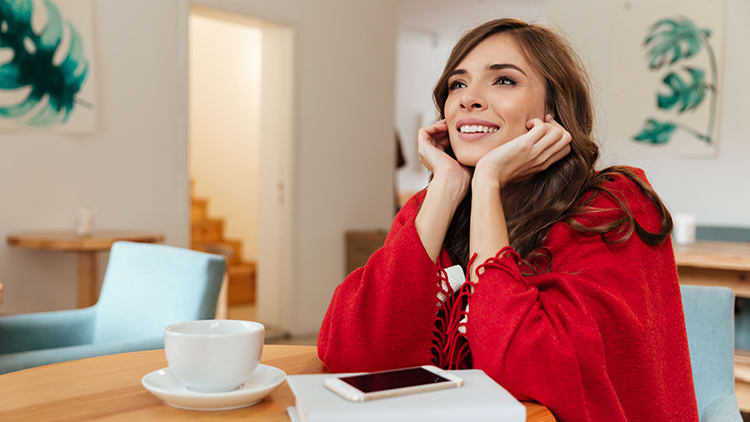Una mujer con una camiseta roja, sentada en una mesa con una taza de café y un teléfono, sonriendo y mirando hacia arriba.