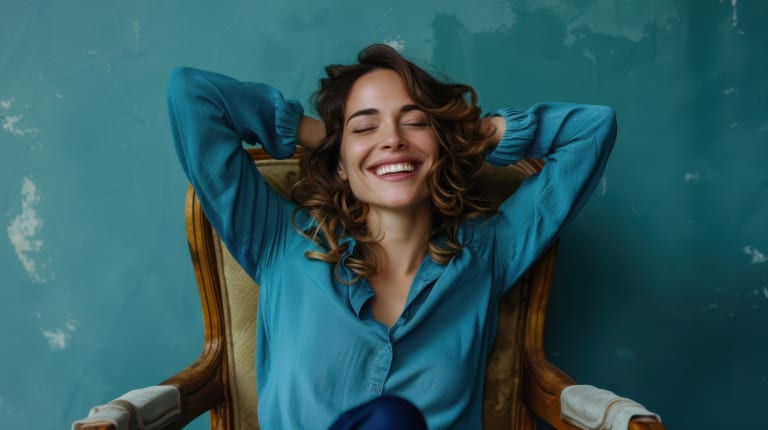 Mujer sonriente con camisa azul se relaja en una silla con las manos detrás de la cabeza contra una pared verde azulado.