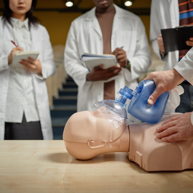 cpr mannequin with a breathing mask on a table and three individuals in white healthcare jackets taking notes