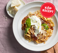 En vit tallrik med pasta med röd arrabiatasås, burrata, parmesan och basilika ligger tillsammans med en liten vit tallrik med parmesan och en orange tygservett på en rosa tygduk.