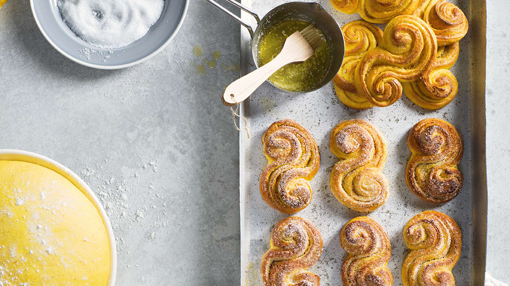 En plåt med saffransbullar bredvid en tallrik med strösocker, en vit skål med gul deg och en metallskål med smält smör och en pensel. 