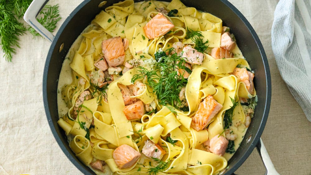 Krämig Laxpasta med babyspenat, lök, vitlök, dill och gräslök i en svart kastrull på en bordsduk.