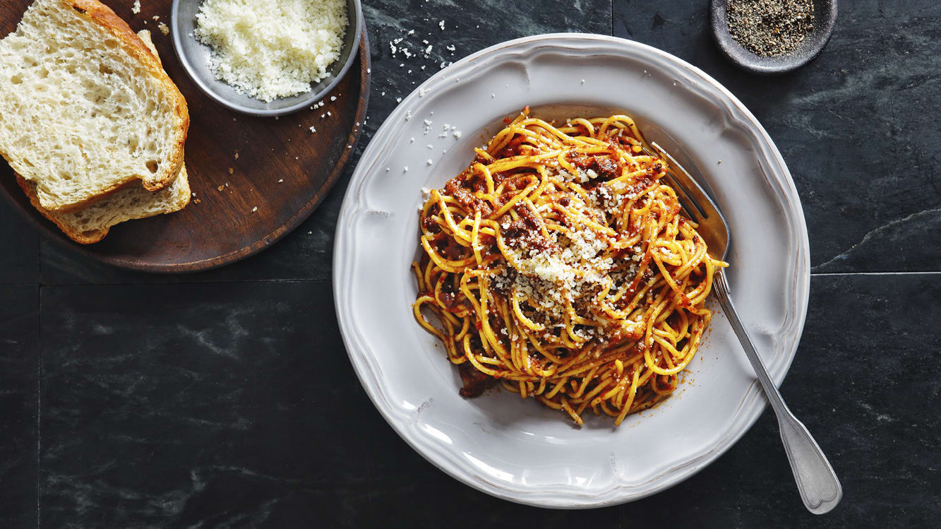 Spaghetti med köttfärssås toppad med riven parmesan på en vit tallrik på ett marmorbord.