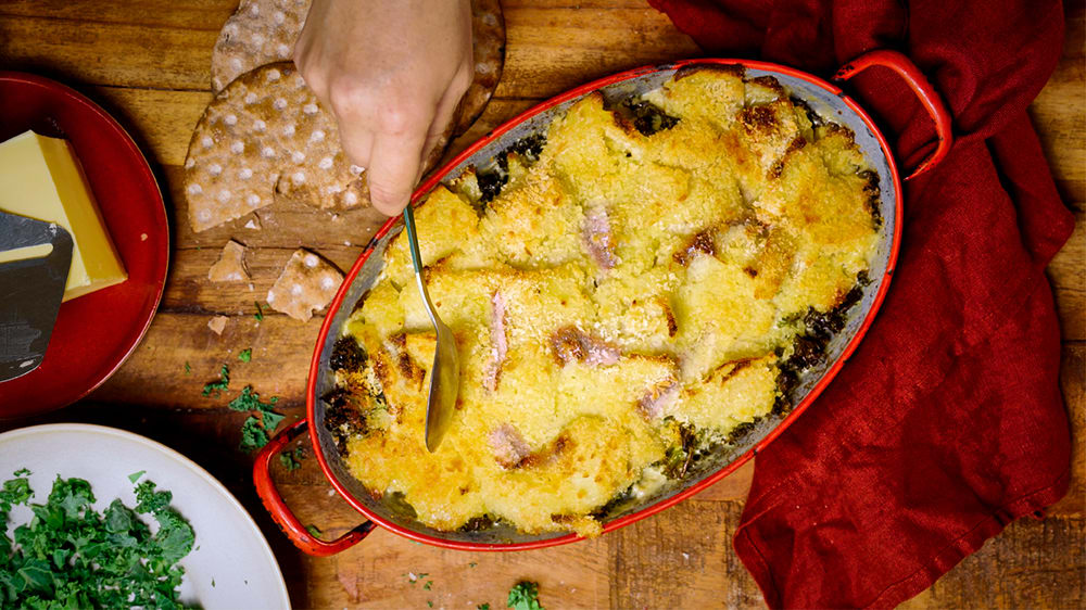 Ett bord med en ugnsform gratinerad Jansson med ansjovis, en tallrik med strimlad grönkål, en tallrik med hårdost och en osthyvel på samt bitar av knäckebröd runtom.