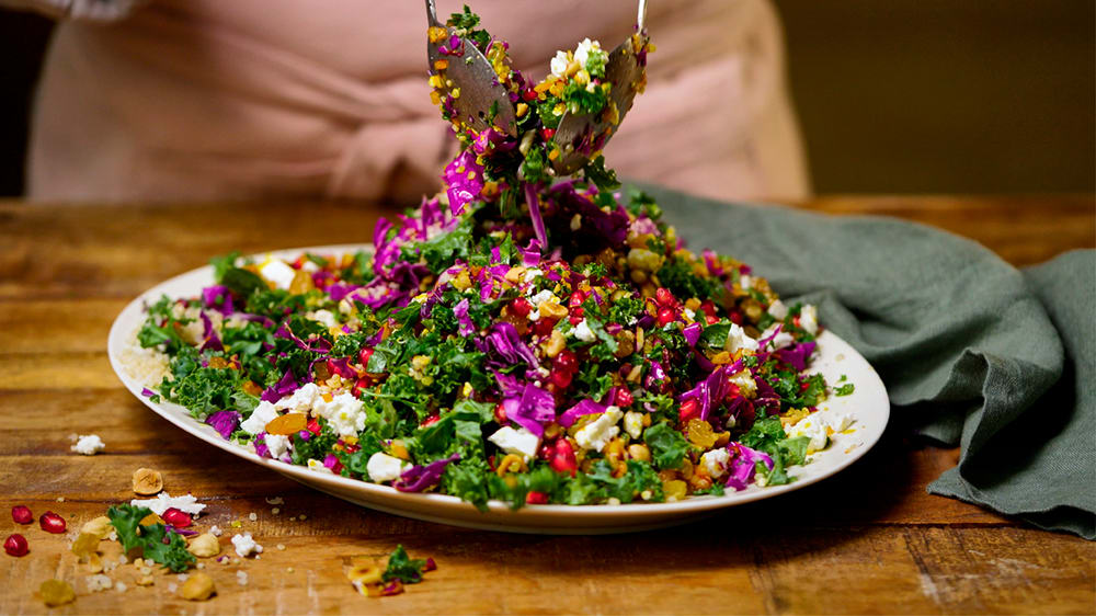 En person i bakgrunden blandar runt en färgglad sallad med ett par salladsbestick på en tallrik innehållandes bland annat finhackad grönkål, hackade nötter, granatäppelkärnor och smulad fetaost.