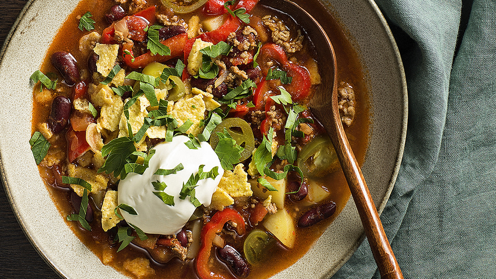 En djup tallrik med en träsked i en soppa med stekt nötfärs, strimlad paprika, kidneybönor, krossade tortillachips, skivad jalapeno och hackad koriander.
