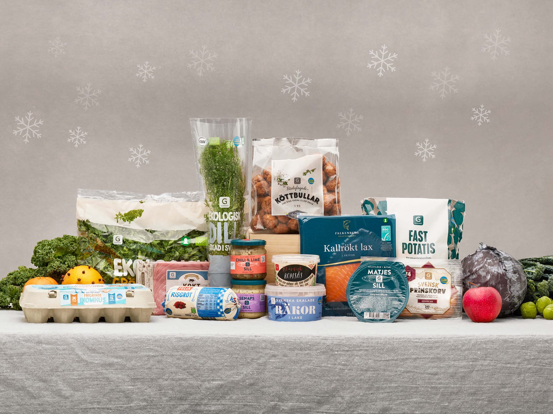 Olika matvaror till julbordet som färdiga köttbullar, matjsessill, ägg, färsk fill, gravad lax, potatis m.m. står grupperade på ett bord och omges av stora snöflingor i bakgrunden.