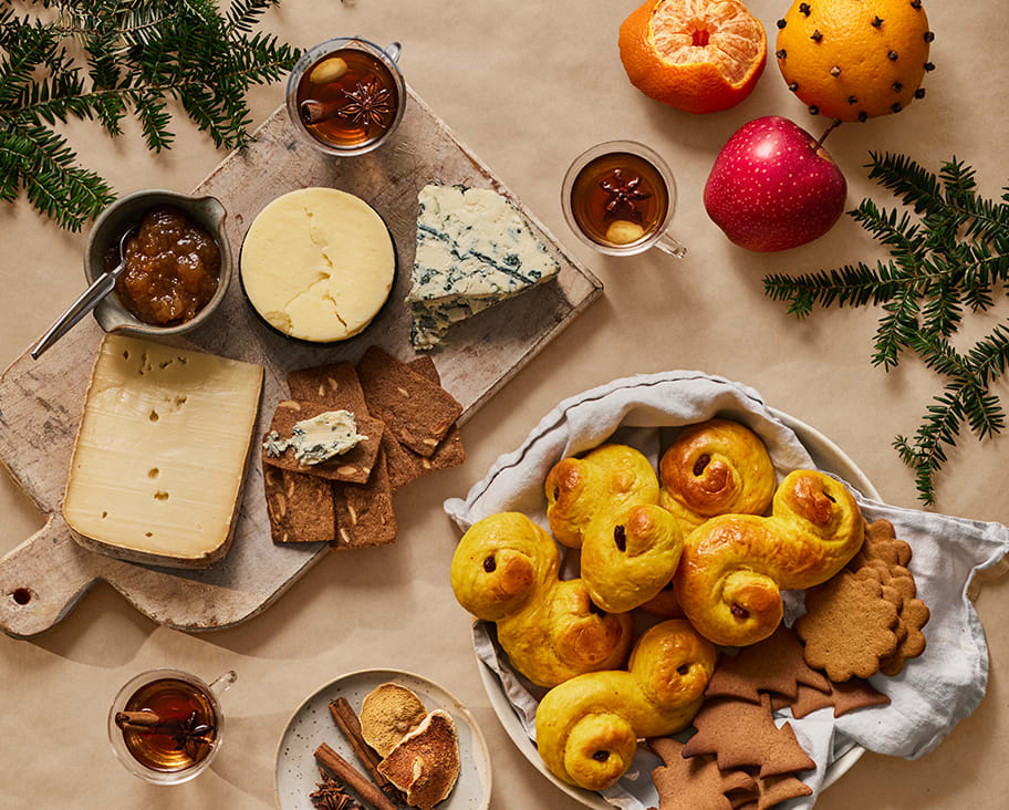 Fotat ovanifrån på ett bord med brun papp ligger lussekatter och pepparkakor, en skärbräda i trä med olika ostar och  en skål med fikonmarmelad, en kökshandduk, en skål med russin och mandlar, två clementiner, tre äpplen och två glas med glögg.