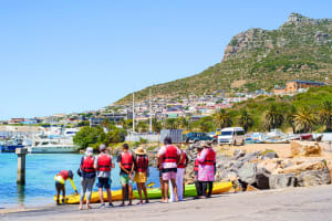 Photo of a tour group wearing red life-jackets standing on the slip way as the tour guide prepares their kayaks for entering the water. In the background are sail boats and a mountain.