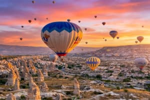 Numerous colorful hot air balloons flying over the unique rock formations and town of Cappadocia, Turkey, during a vibrant sunrise with orange and pink sky.