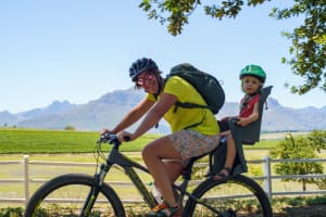 Photo of a lady on a mountain bike with a toddler sitting behind her in a child's seat. In the background are views of the vineyards.