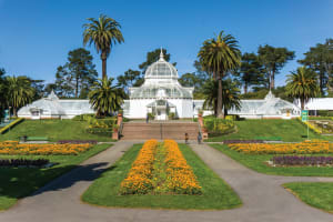 White Victorian conservatory with glass dome surrounded by manicured gardens, palm trees, and colorful flower beds