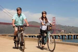 Two cyclists with helmets and bike bags pose in front of the Golden Gate Bridge in San Francisco