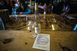 Wine tasting setup at a winery with a bottle, glass, and menu on a wooden table with guests in background