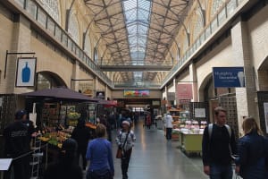 Interior of a bustling covered market hall with vendor stalls, shoppers, and a glass ceiling skylight