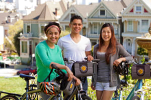 Three young adults with bikes and helmets smiling in front of Victorian houses in San Francisco