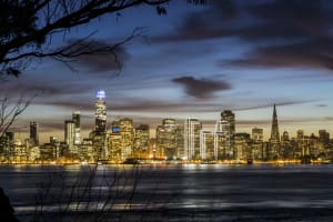 San Francisco skyline at dusk with illuminated buildings reflected in bay waters, framed by bare tree branches
