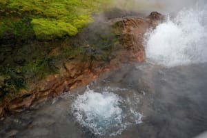 Close aerial view of Deildartunguhver geothermal hot spring, with boiling water splashing over rust-colored volcanic rock surrounded by vivi