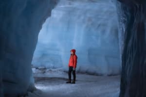 A lone visitor in a red jacket and hood standing inside a large chamber within the Langjokull glacier ice tunnel system, surrounded by tower