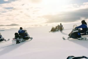 Several snowmobilers in blue suits riding across the wide flat expanse of Langjokull glacier toward a bright overcast horizon, with a guide 