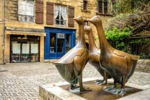 Bronze statue of three geese in a small square in Sarlat-la-Canéda, with stone façades and a blue shopfront behind